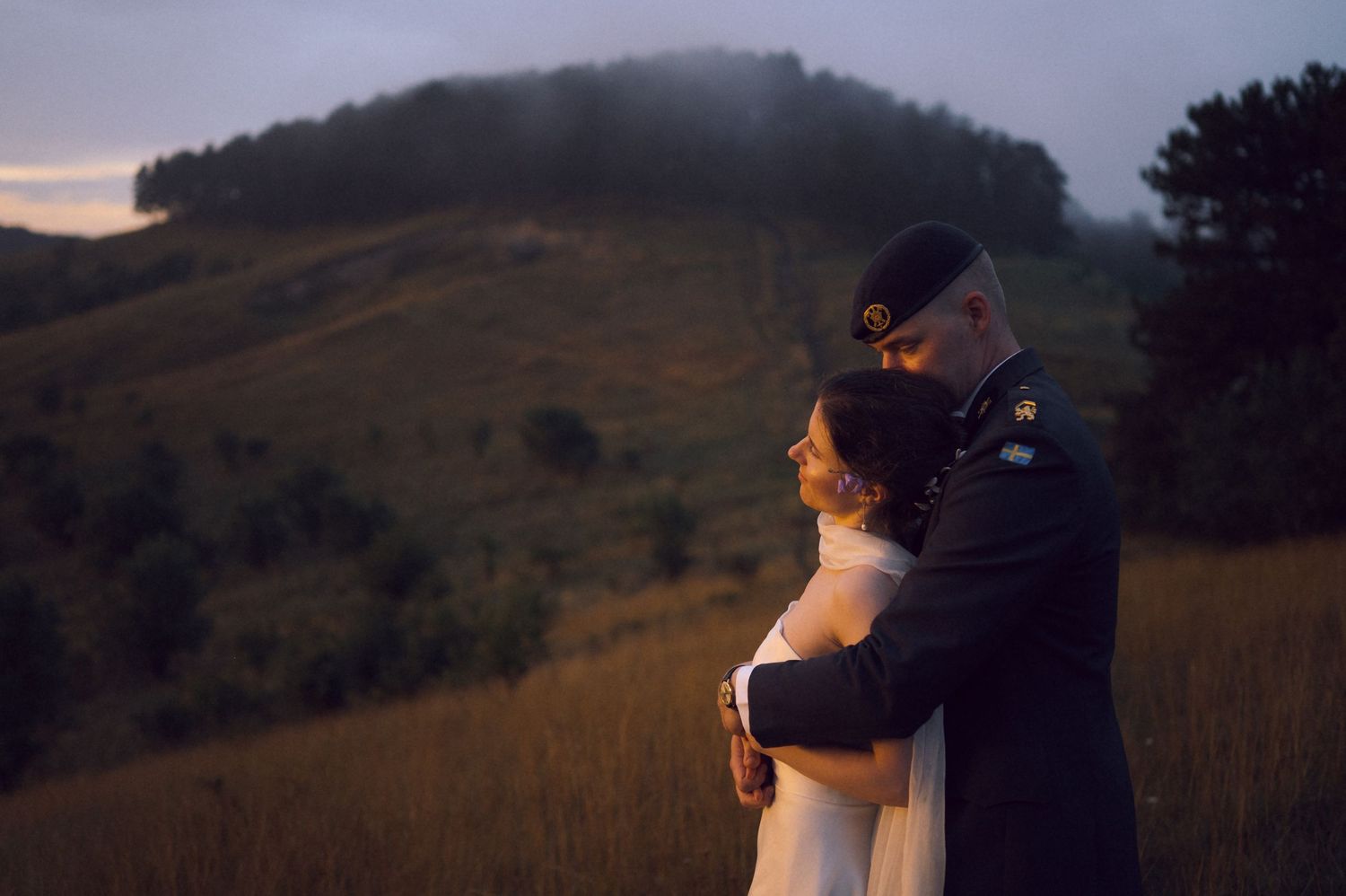 Military groom in uniform embraces bride in white dress at golden hour with misty green hills in background.