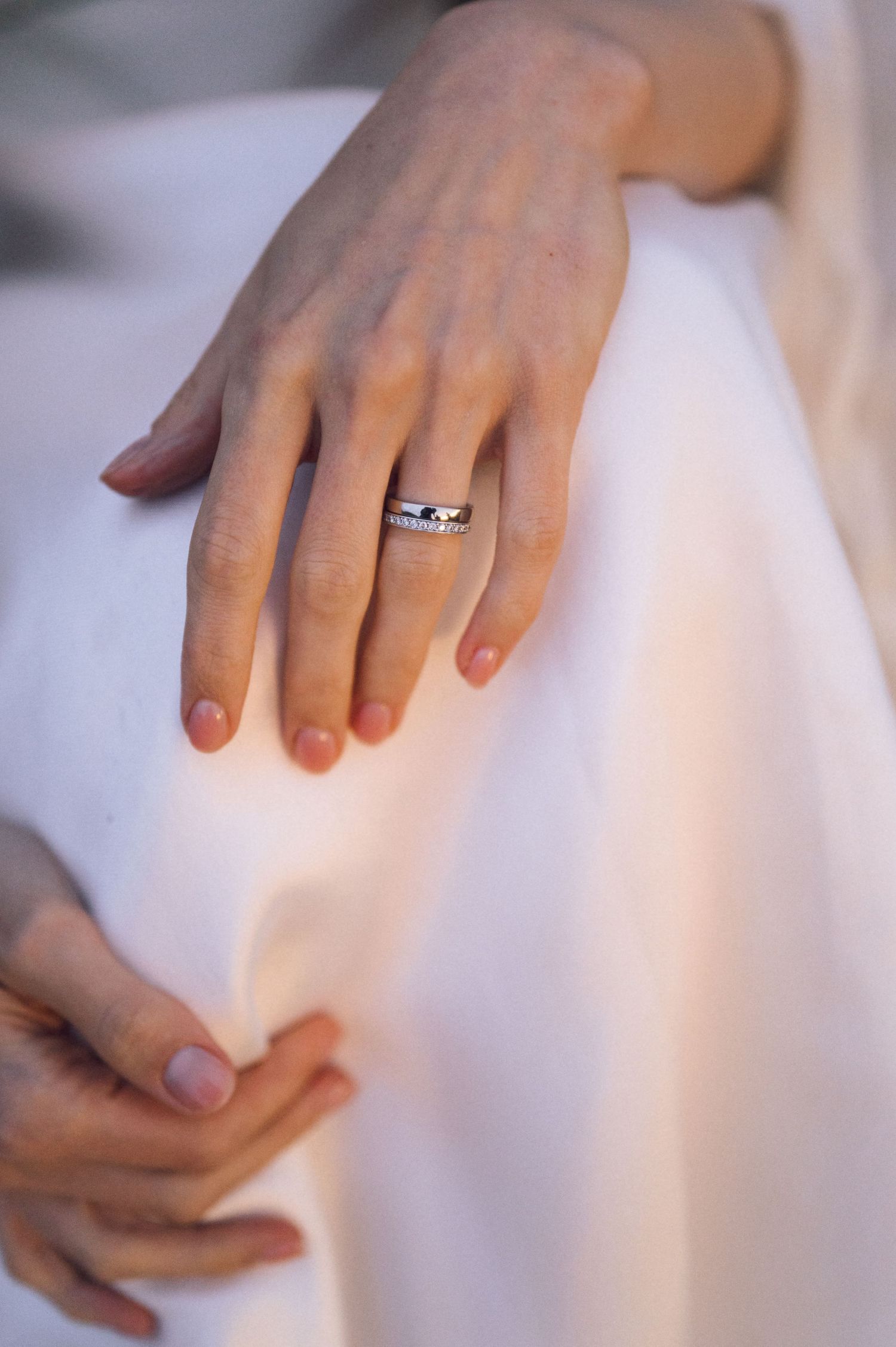 Close-up of a woman's hands wearing a silver diamond ring on her finger, resting on a white dress.