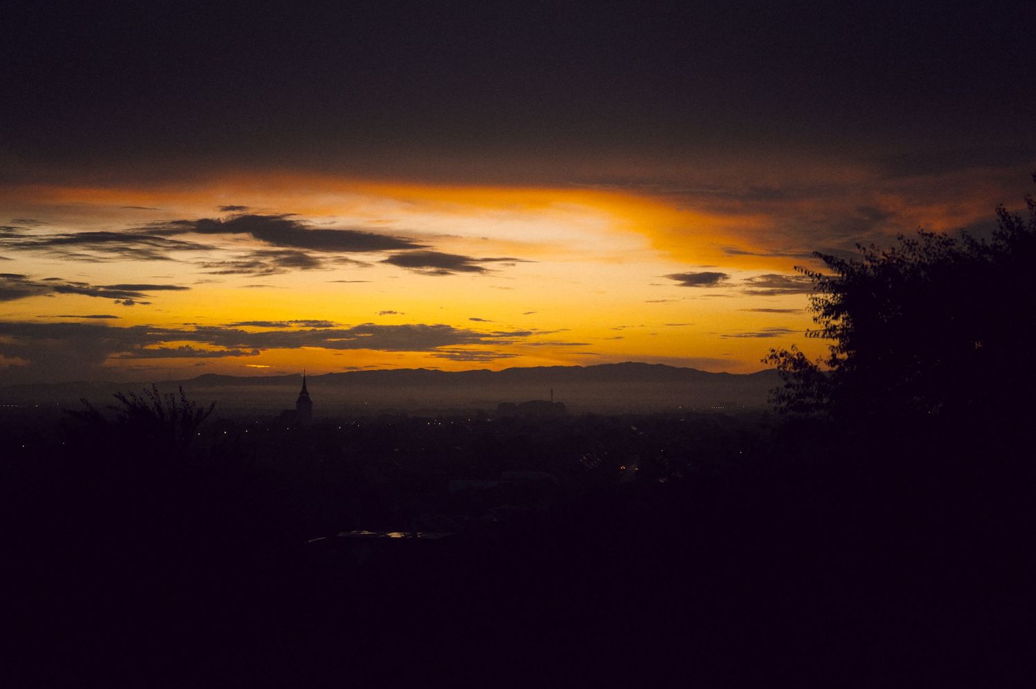 Dramatic sunset skyline silhouette with vibrant orange and golden sky over a distant cityscape and rolling hills.