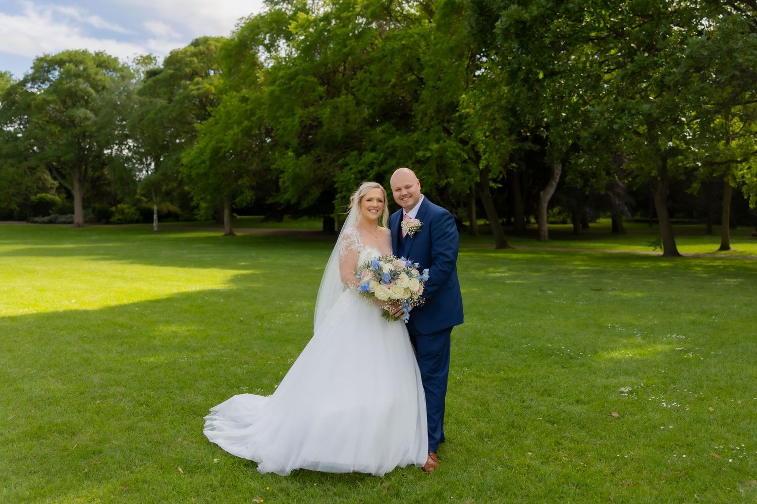 Bride & groom posing together on lush green grass, bride holding bouquet in white gown, groom in navy suit. The Grand Hotel, Casual Captures