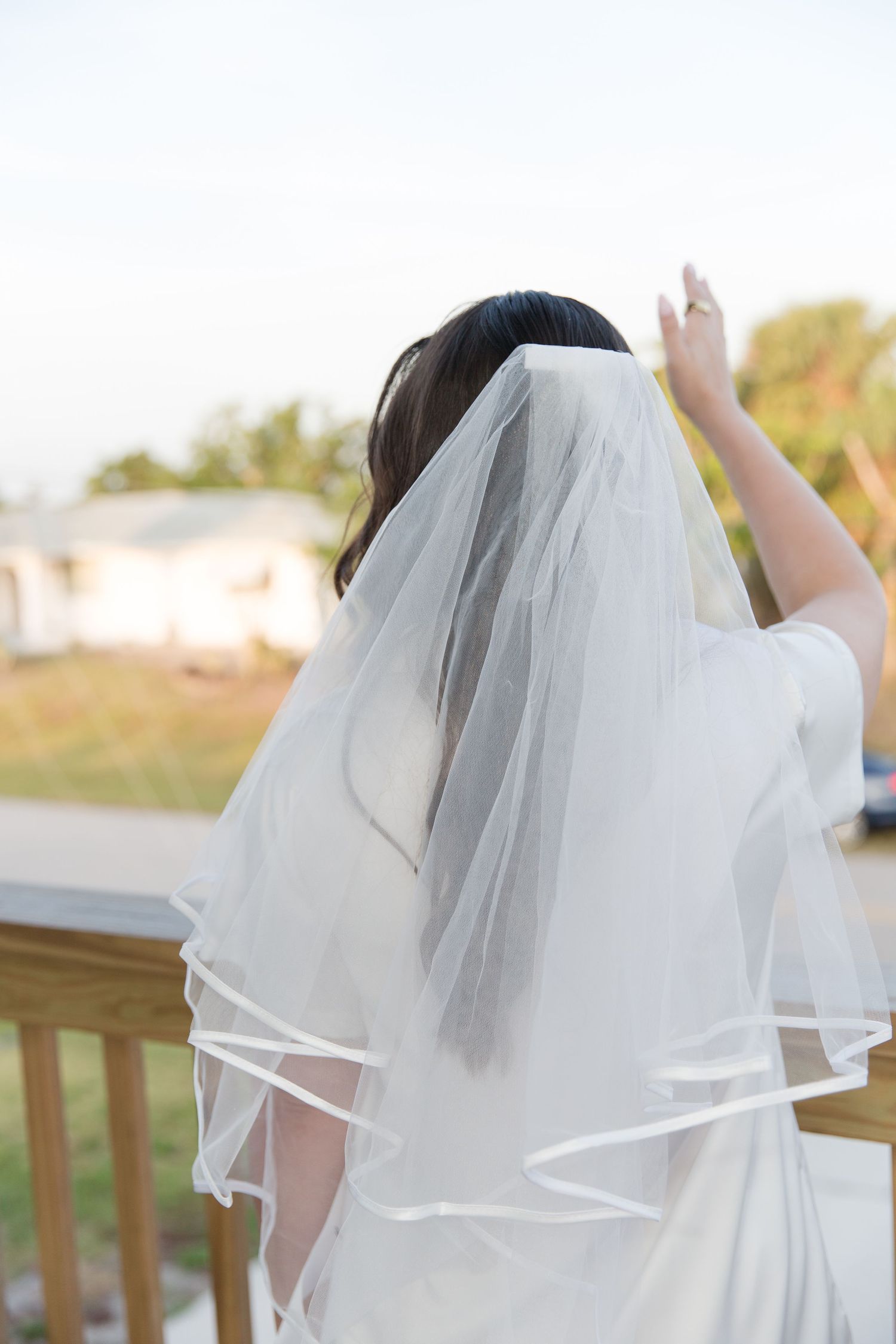 Bride in white wedding dress and two-tier veil standing on wooden deck, viewed from behind.