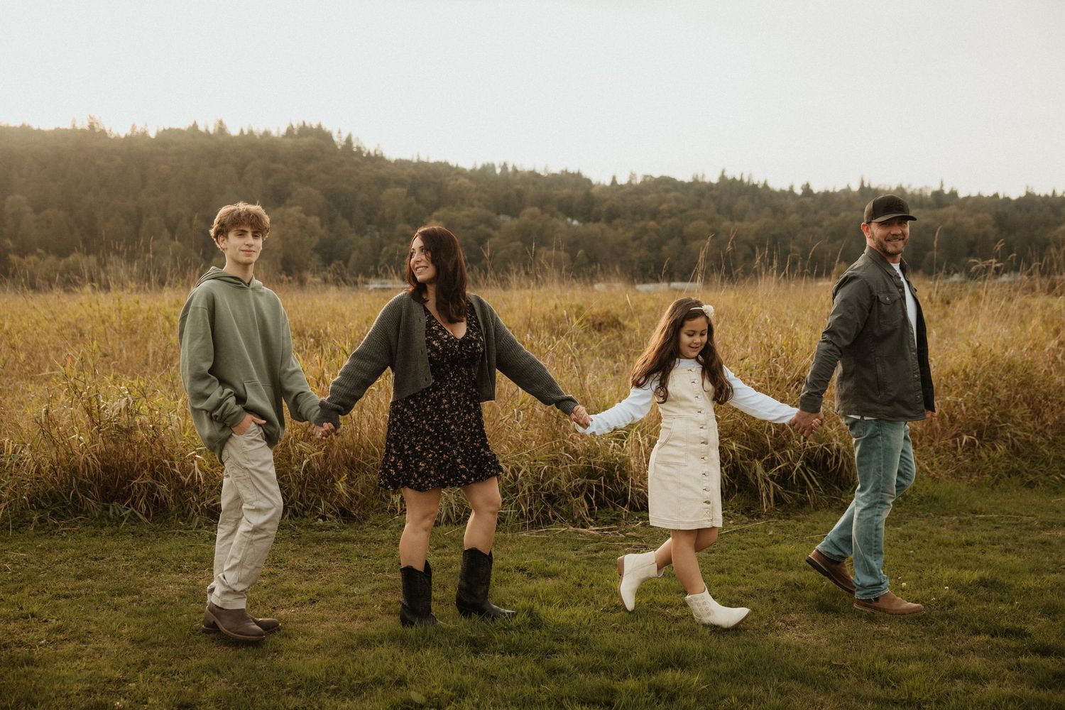 Family of four holding hands walking through golden autumn field with forested hills in background.