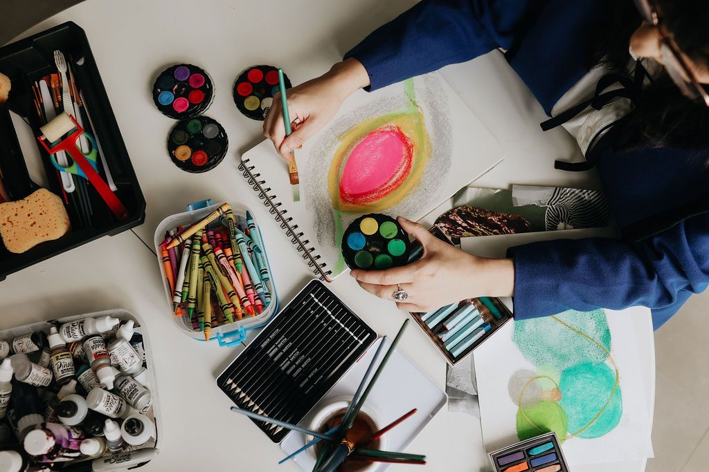 Overhead view of an artist painting with watercolors, surrounded by crayons, brushes, and art supplies on a white desk.