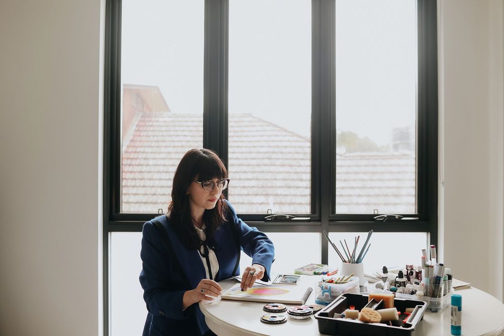 Woman in blue blazer applying makeup at a desk by a large window with natural light.