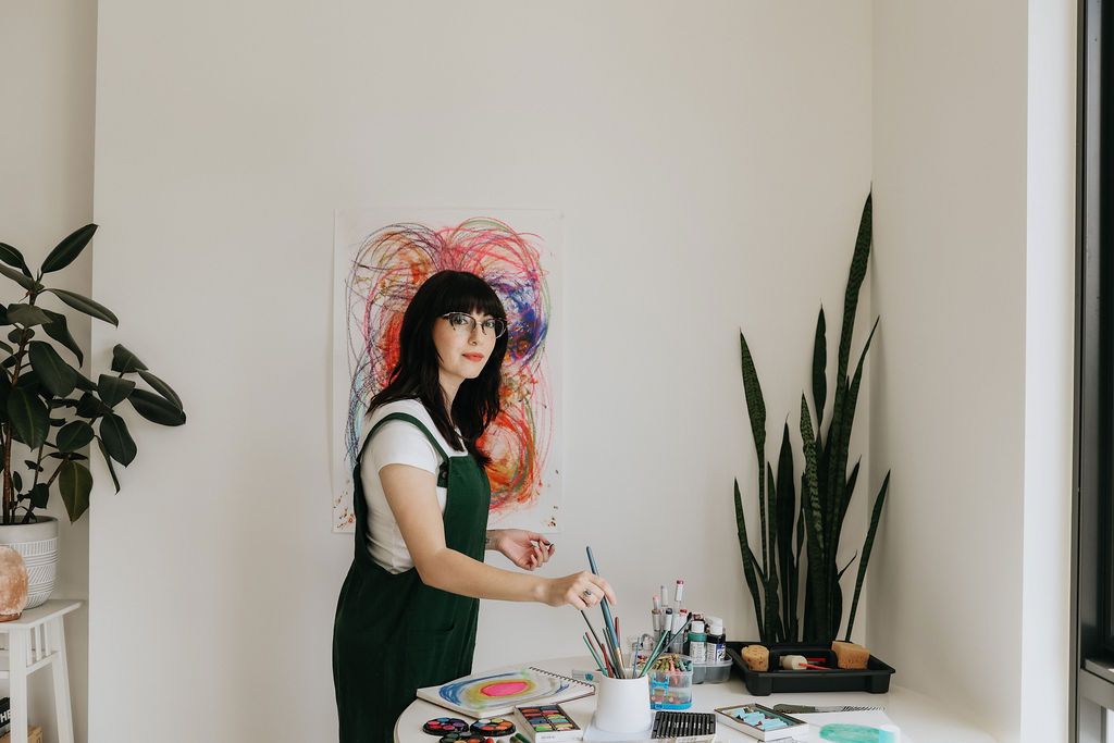 Artist in green overalls painting at a white table with colorful abstract artwork on wall in bright studio.