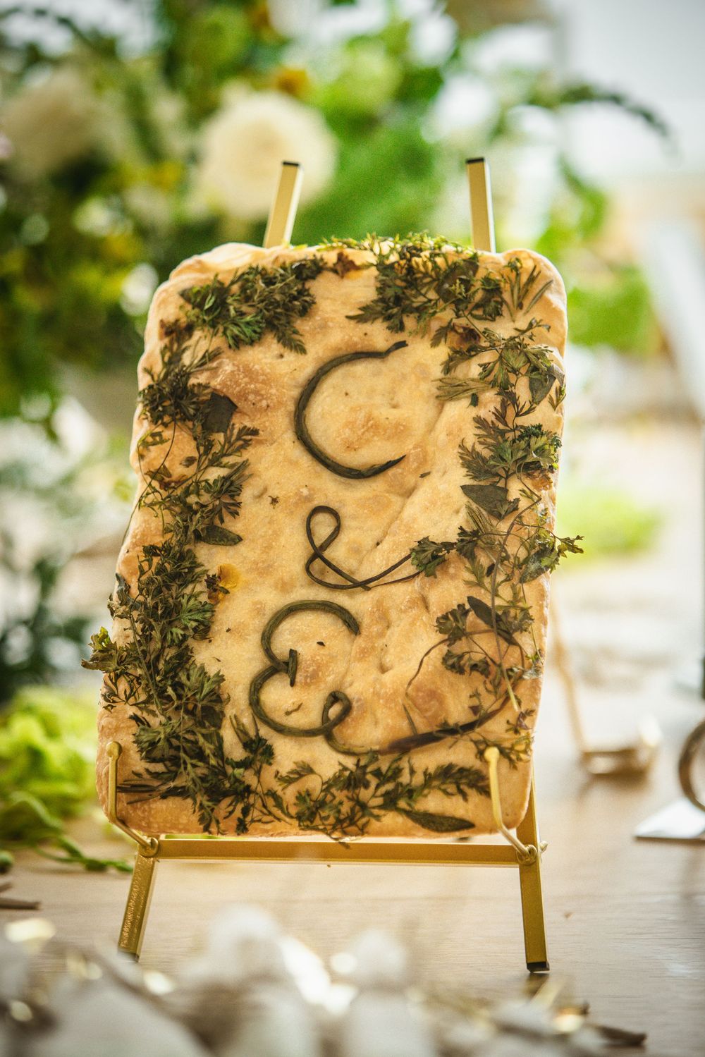 Focaccia bread wedding sign with herb garnish and couple's initials displayed on a gold easel stand.
