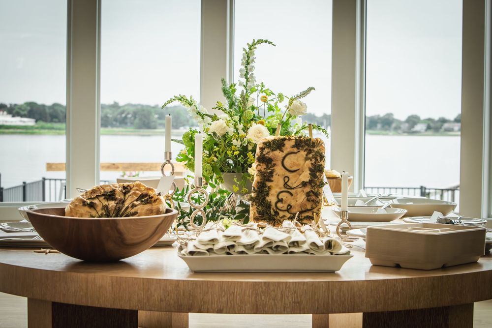 Elegant wedding reception table with floral centerpiece, wooden bowl of treats, and waterfront view through large windows.