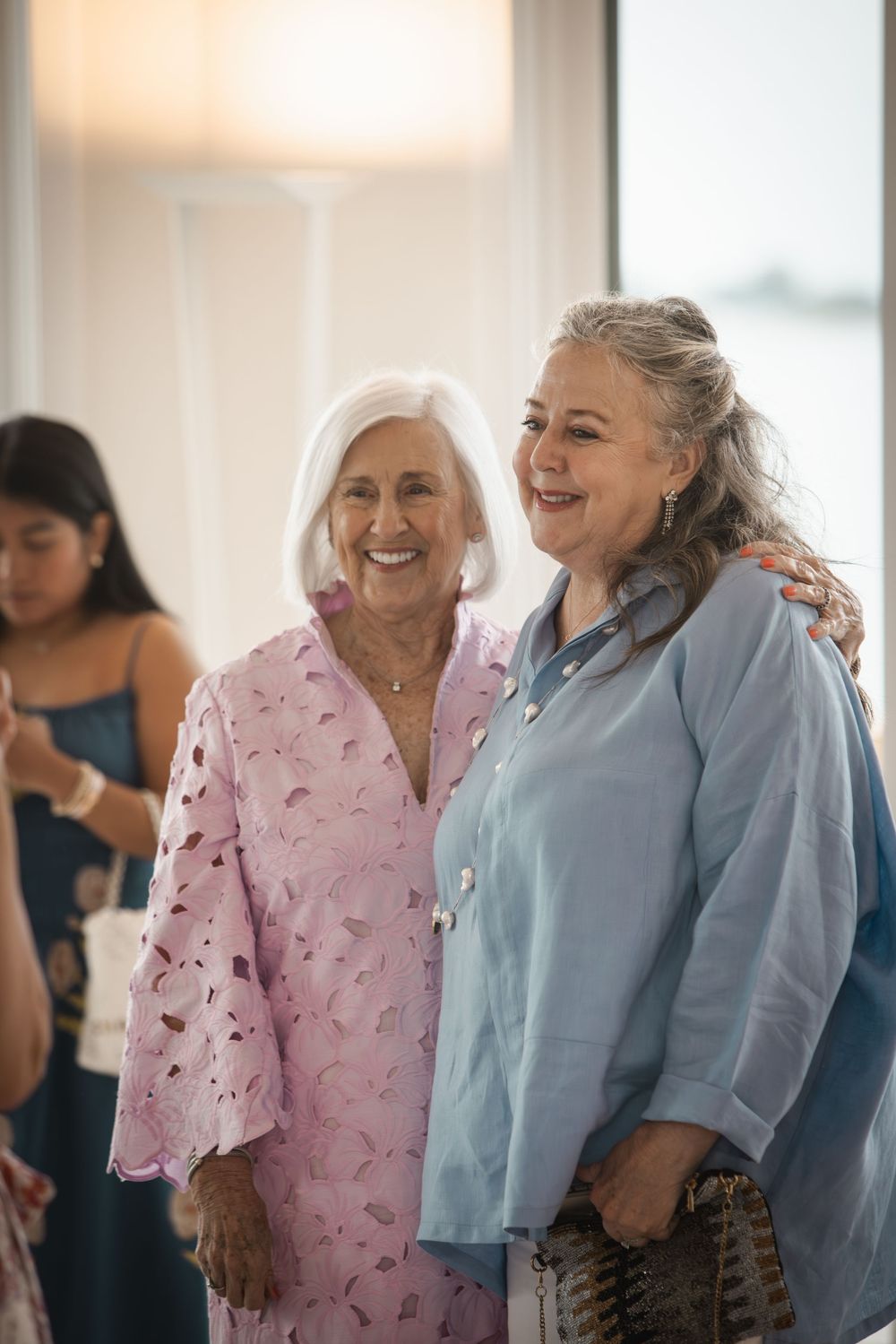 Two smiling older women in pink and blue outfits posing together at a social gathering.