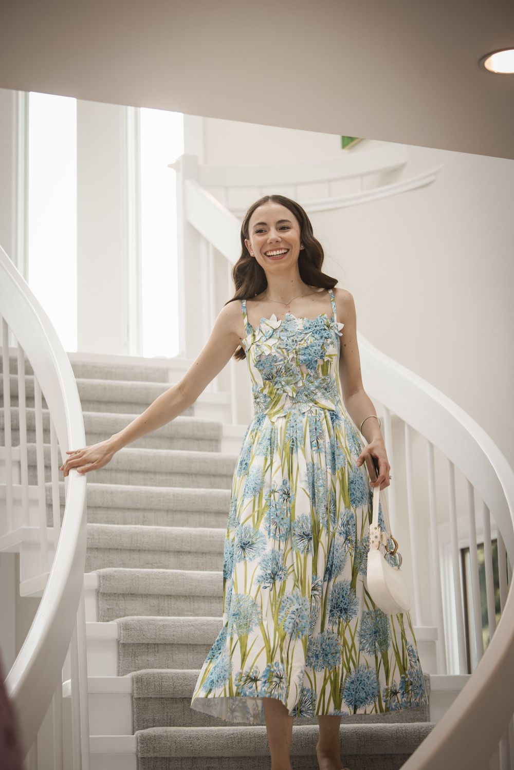 Smiling woman in floral midi dress descending elegant white curved staircase, holding handbag.