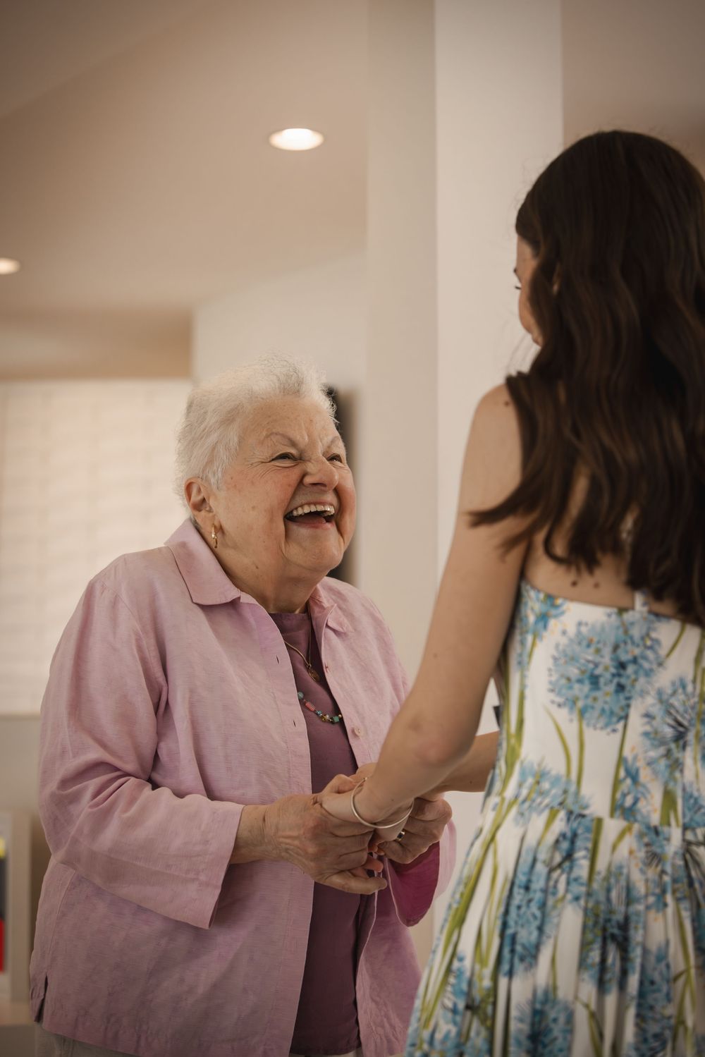 Elderly woman in pink jacket laughs joyfully while holding hands with young brunette woman in floral dress indoors.