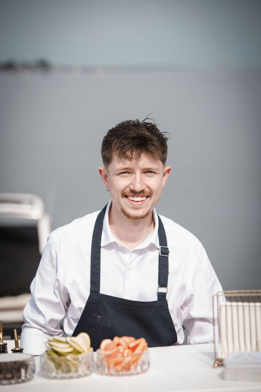 Smiling young male chef in white shirt and black apron standing at outdoor food station near waterfront.
