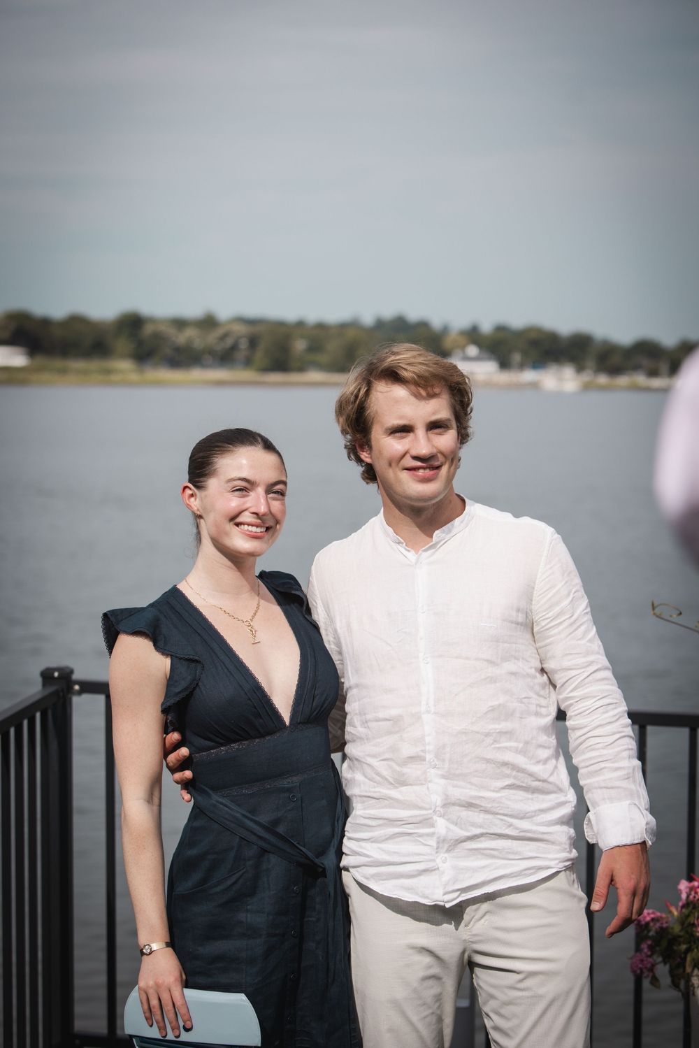 A smiling couple poses outdoors near a waterfront, woman in black dress and man in white shirt and khaki pants.