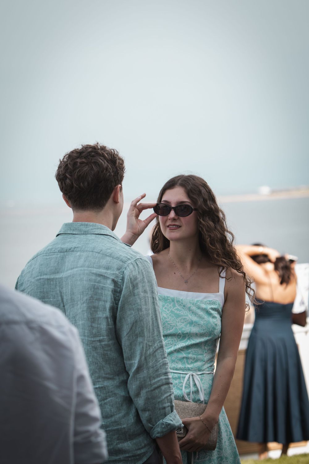 Young couple in matching teal outfits conversing outdoors near water, woman wearing sunglasses smiling at man.