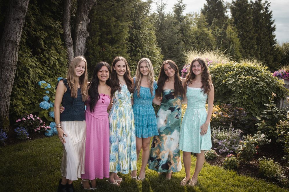 Six young women in colorful summer dresses smiling together in a lush garden setting.