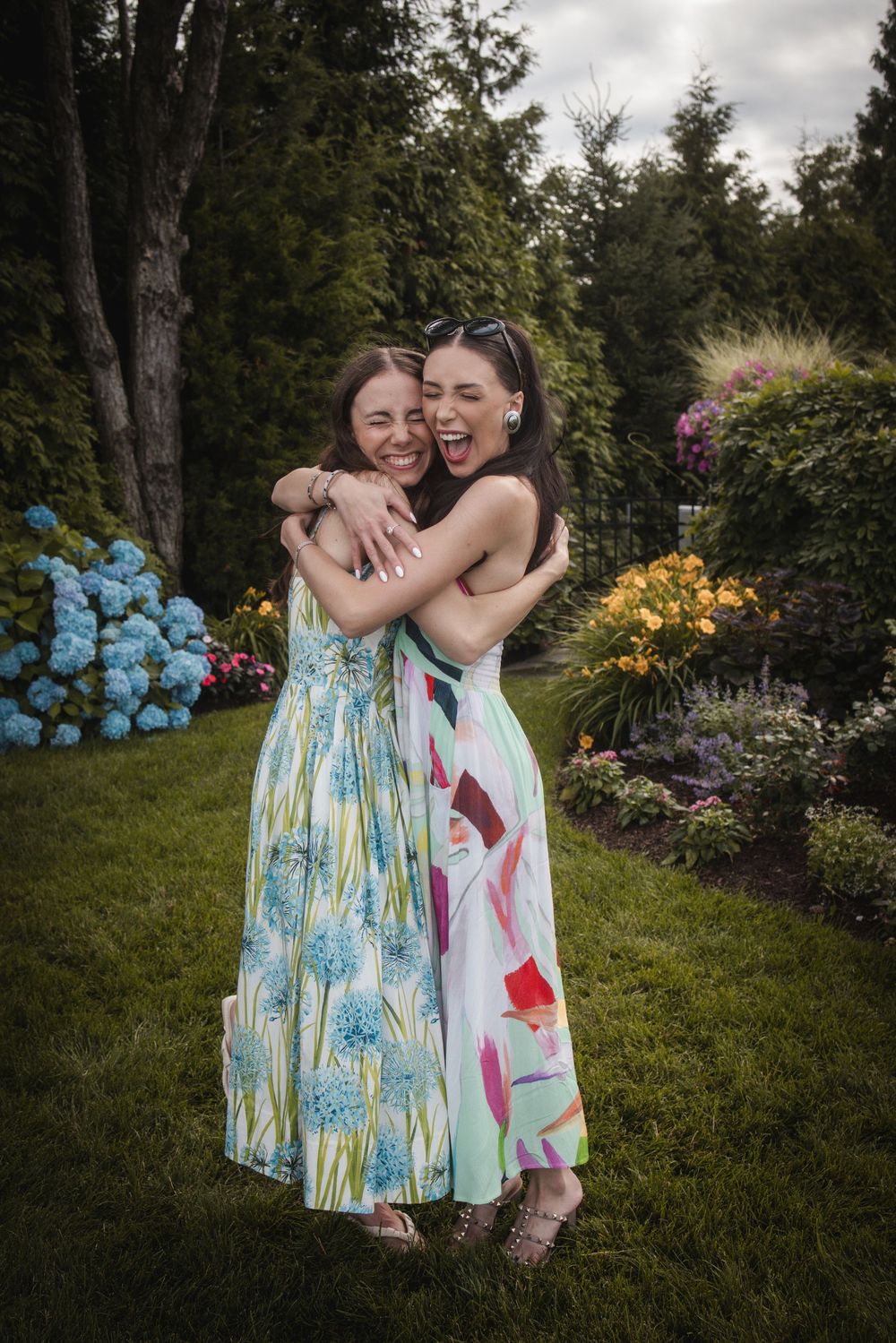 Two women in floral maxi dresses hugging and laughing in a lush garden with colorful hydrangeas.