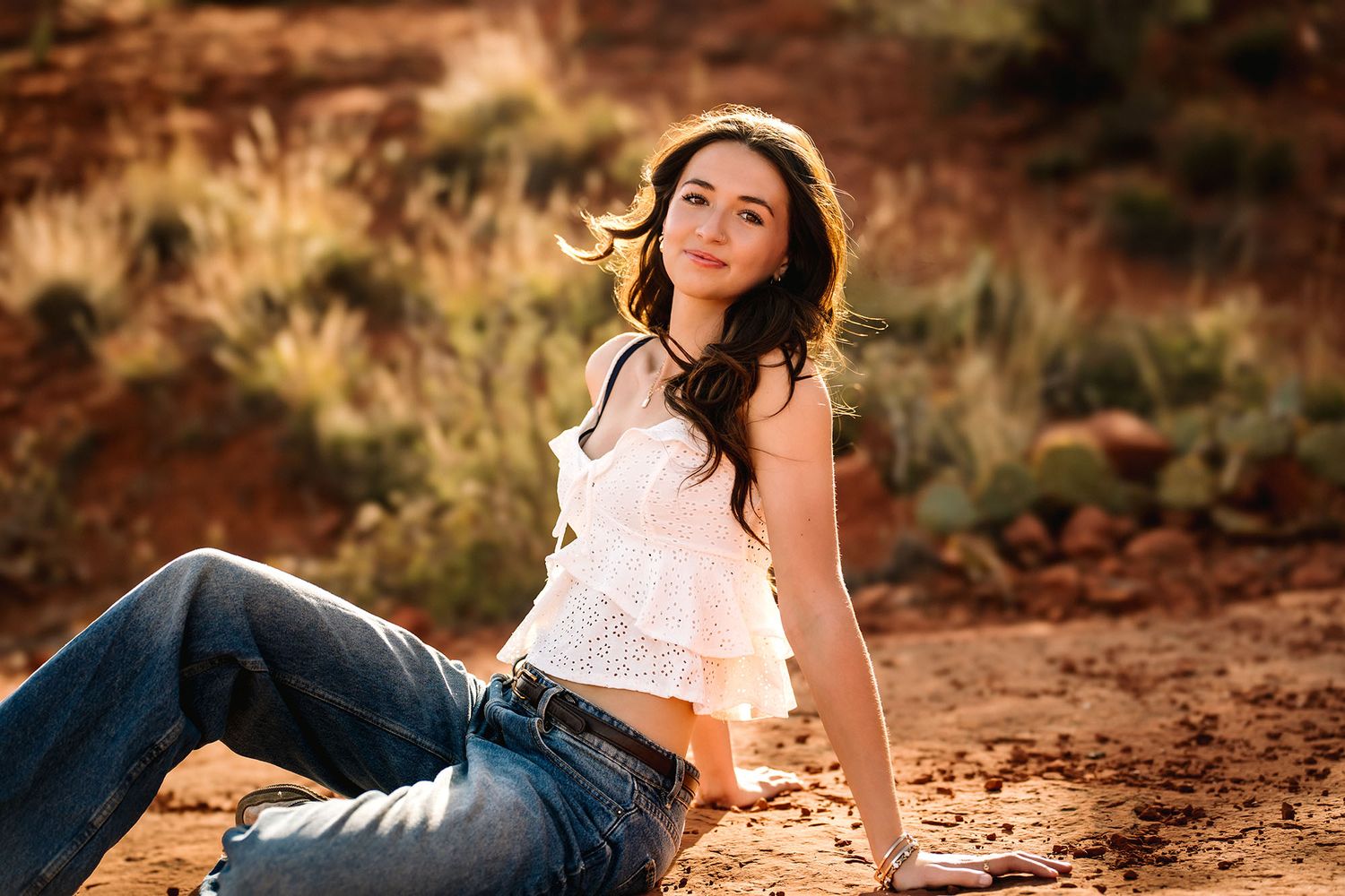 Senior with dark hair sits on red desert sand, wearing white top and jeans, smiling in warm sunlight.