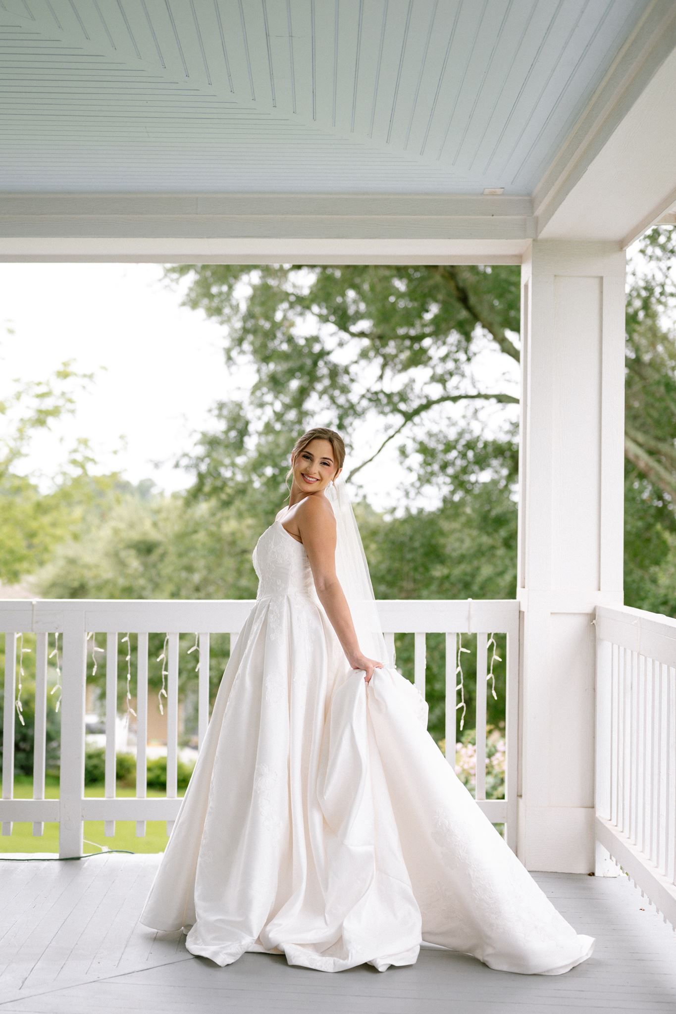 Bride in elegant white ballgown standing on a classic white porch, smiling over her shoulder with lush greenery behind her.