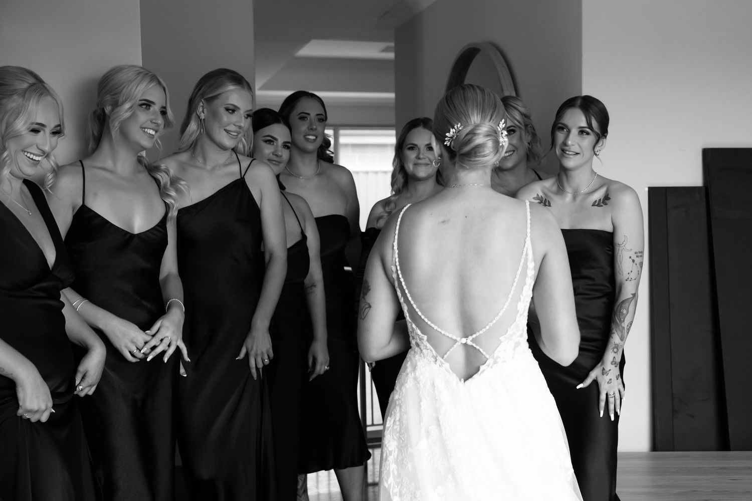 Bride in lace backless gown reveals look to bridesmaids in black dresses in elegant black and white wedding photo.