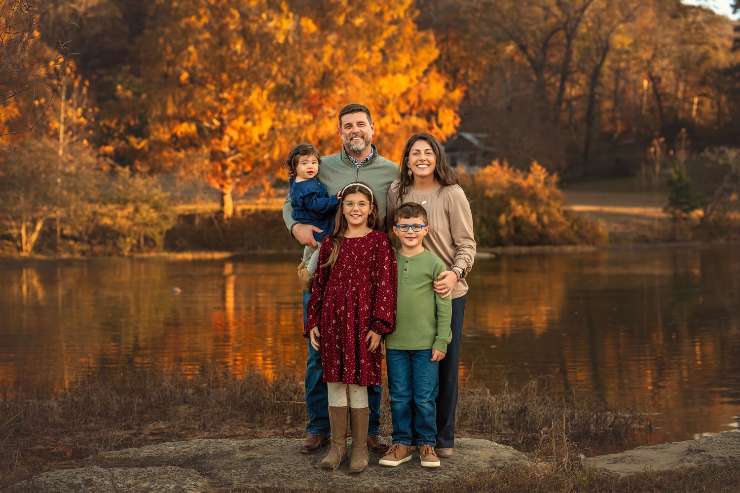 Family of five posing in a sunlit wildflower field at golden hour, parents holding a baby in a lush green meadow.