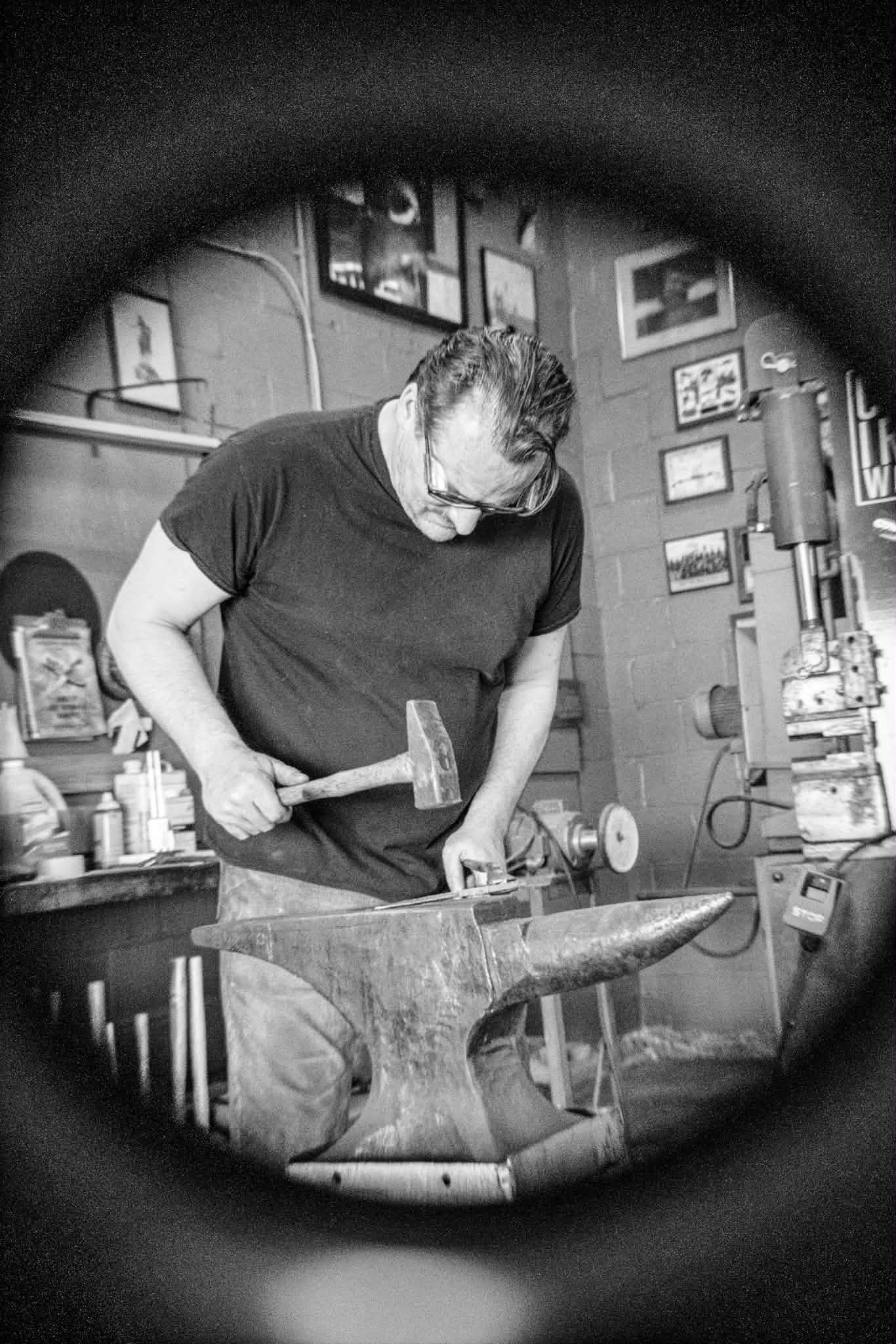 Black and white photo of a blacksmith hammering metal on an anvil in a workshop, viewed through a circular frame.