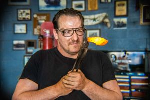 A man in a black t-shirt holds glowing molten glass on a metal rod in a workshop filled with photos.
