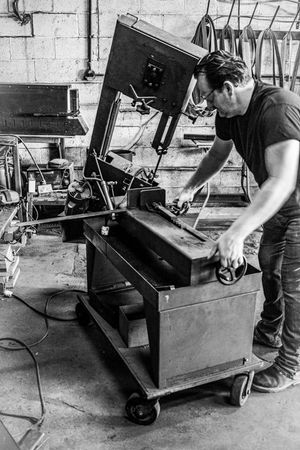 Male metalworker operating a band saw machine in a black and white industrial workshop setting.