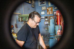 Man in black t-shirt and glasses works in a vintage blue workshop, viewed through a circular frame.