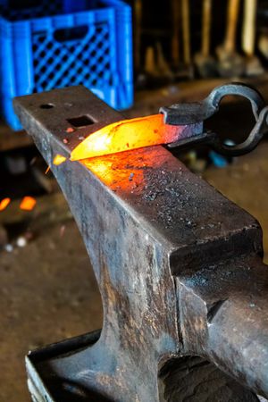 Glowing hot metal piece resting on a blacksmith anvil in a forge workshop with sparks and tools in background.