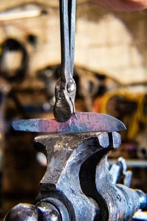 Close-up of a blacksmith forging a metal hook in a workshop vise, with blurred tools in the background.