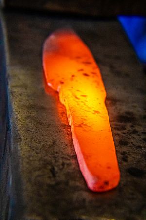 Glowing red-hot metal blade being forged on an anvil in a blacksmith workshop.