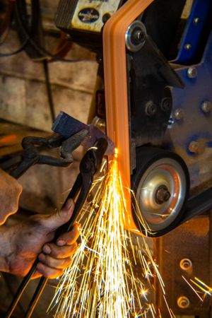 Worker grinding metal on a belt sander with bright orange sparks flying in a workshop.