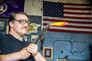Man with glasses holding glowing hot molten glass on metal rod in workshop with American flag background.