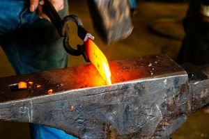 Blacksmith pouring glowing molten metal on an anvil in a dark forge workshop.