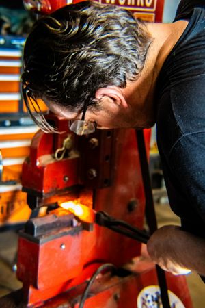 A blacksmith closely inspects glowing hot metal on a red forge anvil in a workshop.