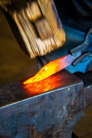 A glowing red-hot metal blade being forged on an anvil with a hammer strike in a blacksmith workshop.
