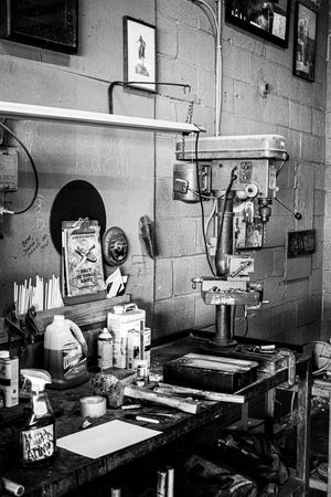 Black and white photo of a cluttered workshop with a drill press, tools, and supplies on a workbench against a cinder block wall.