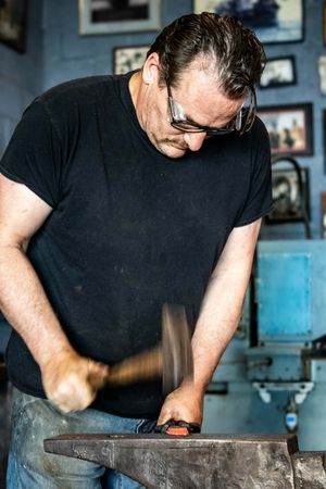 Male blacksmith wearing safety glasses hammering hot metal on an anvil in a workshop.