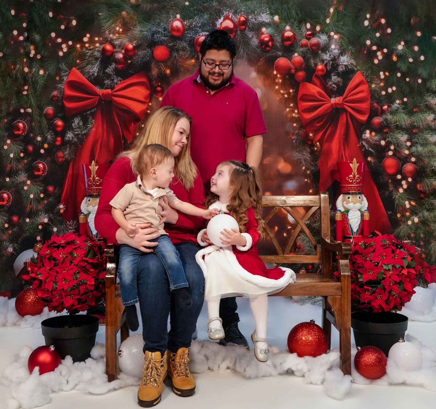 Family of four posing for a festive Christmas photo with holiday decorations, red bows, and poinsettias.