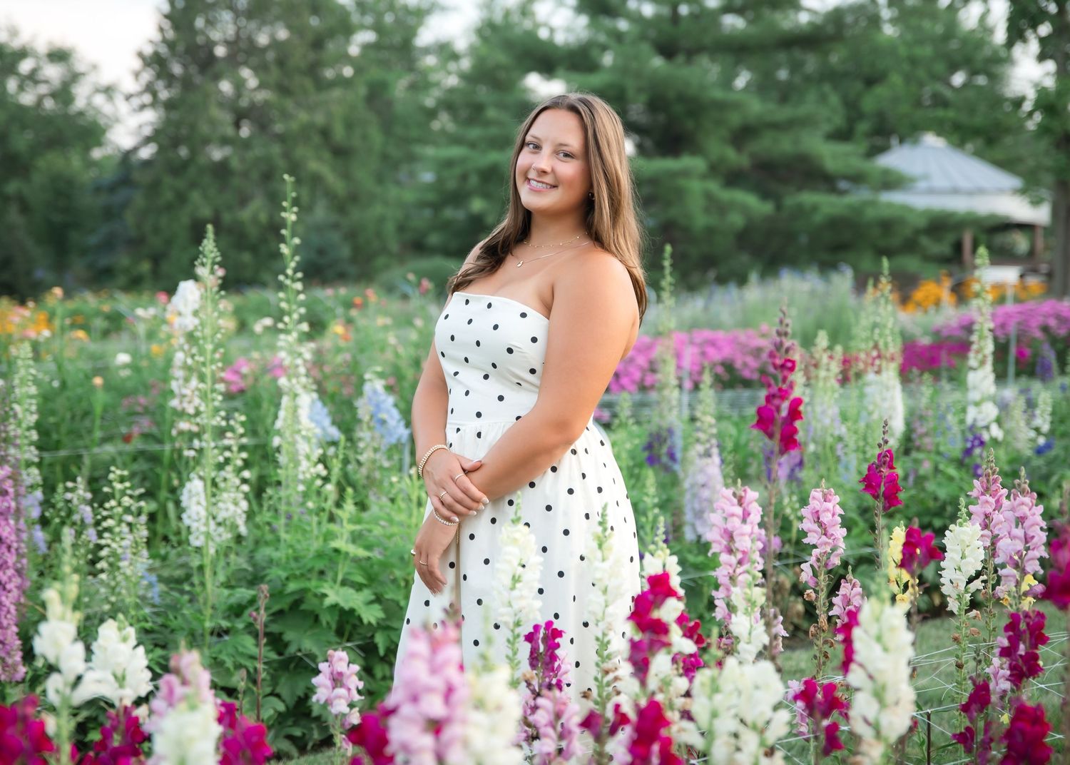 Smiling young woman in white polka dot dress posing among colorful garden flowers including pink and white blooms.