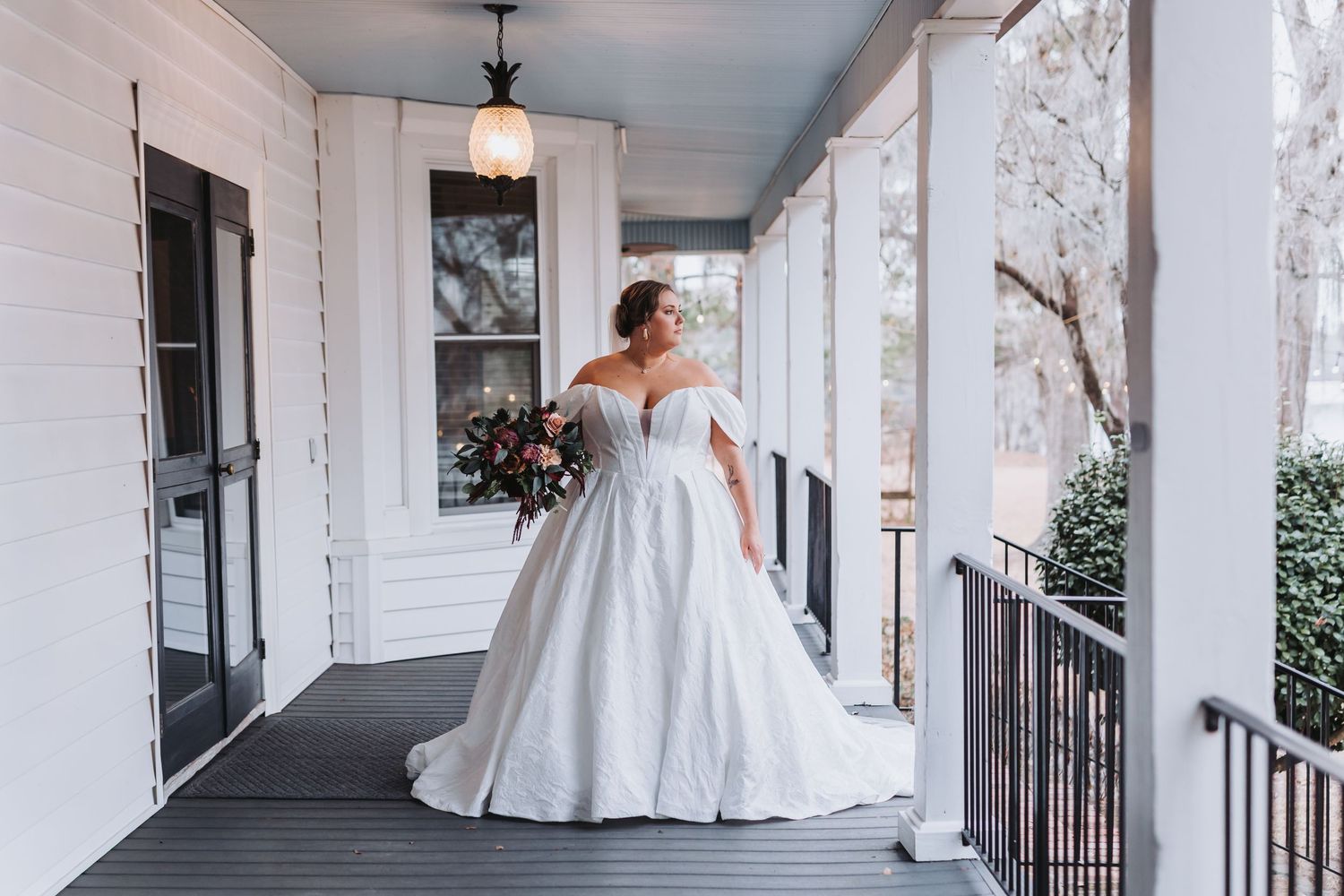 Bride in white lace gown and veil holding colorful bouquet of pink and peach flowers in lush garden setting.