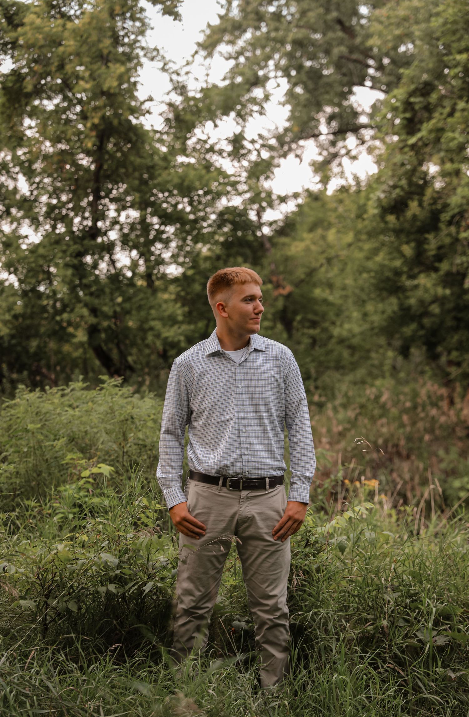 Young man in plaid shirt and khaki pants standing in lush green forest clearing, looking to the side.
