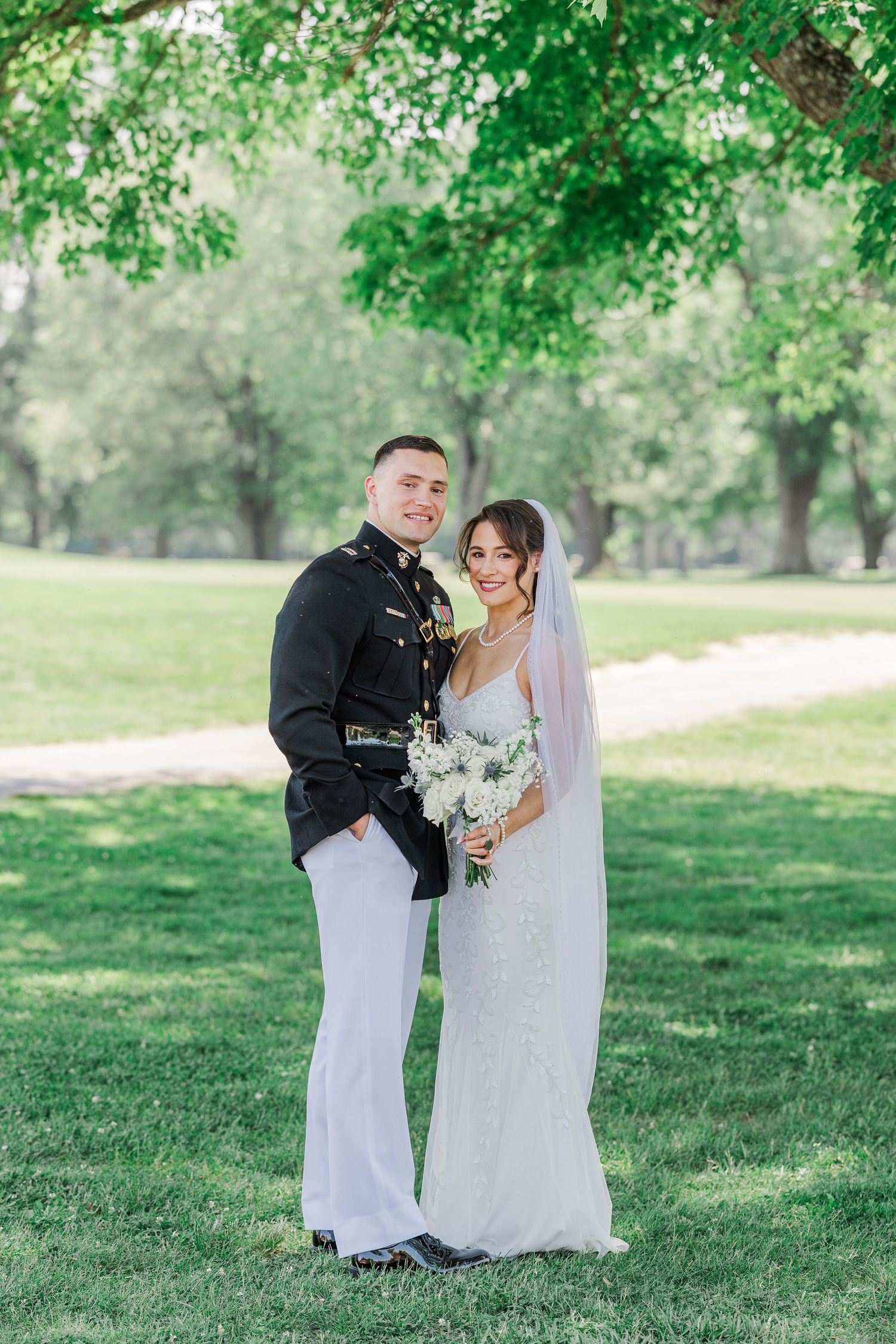 Military groom in dress uniform and bride in white gown holding bouquet pose together outdoors under green trees.