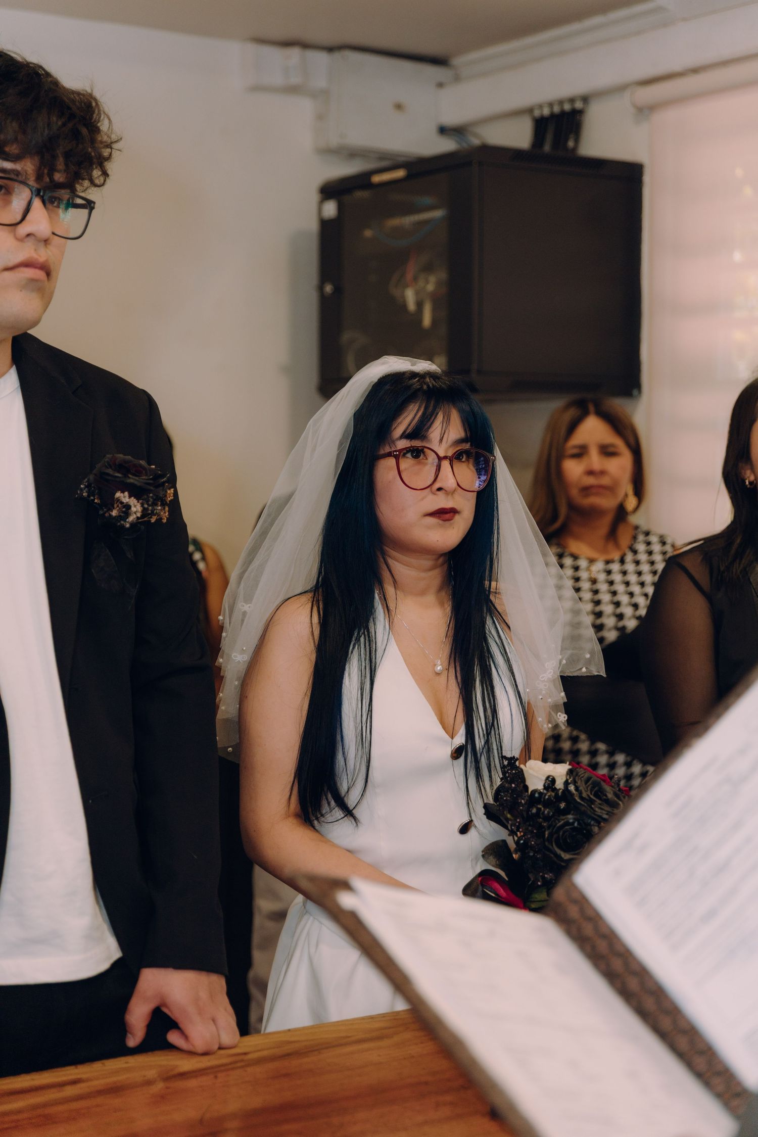 Bride wearing veil and glasses listens attentively during wedding ceremony at wooden altar.