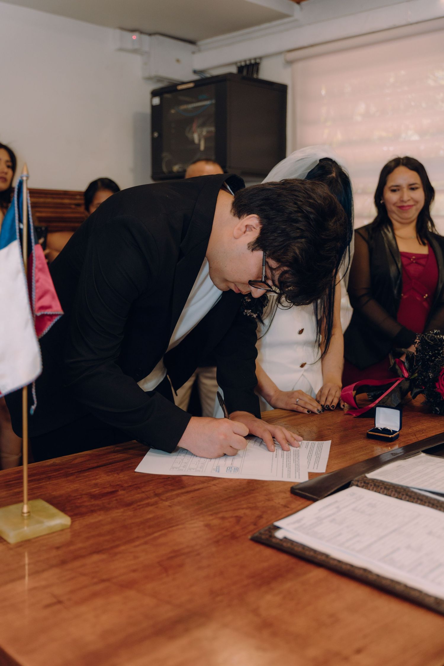 Groom in black suit signing wedding documents at wooden table during civil ceremony.