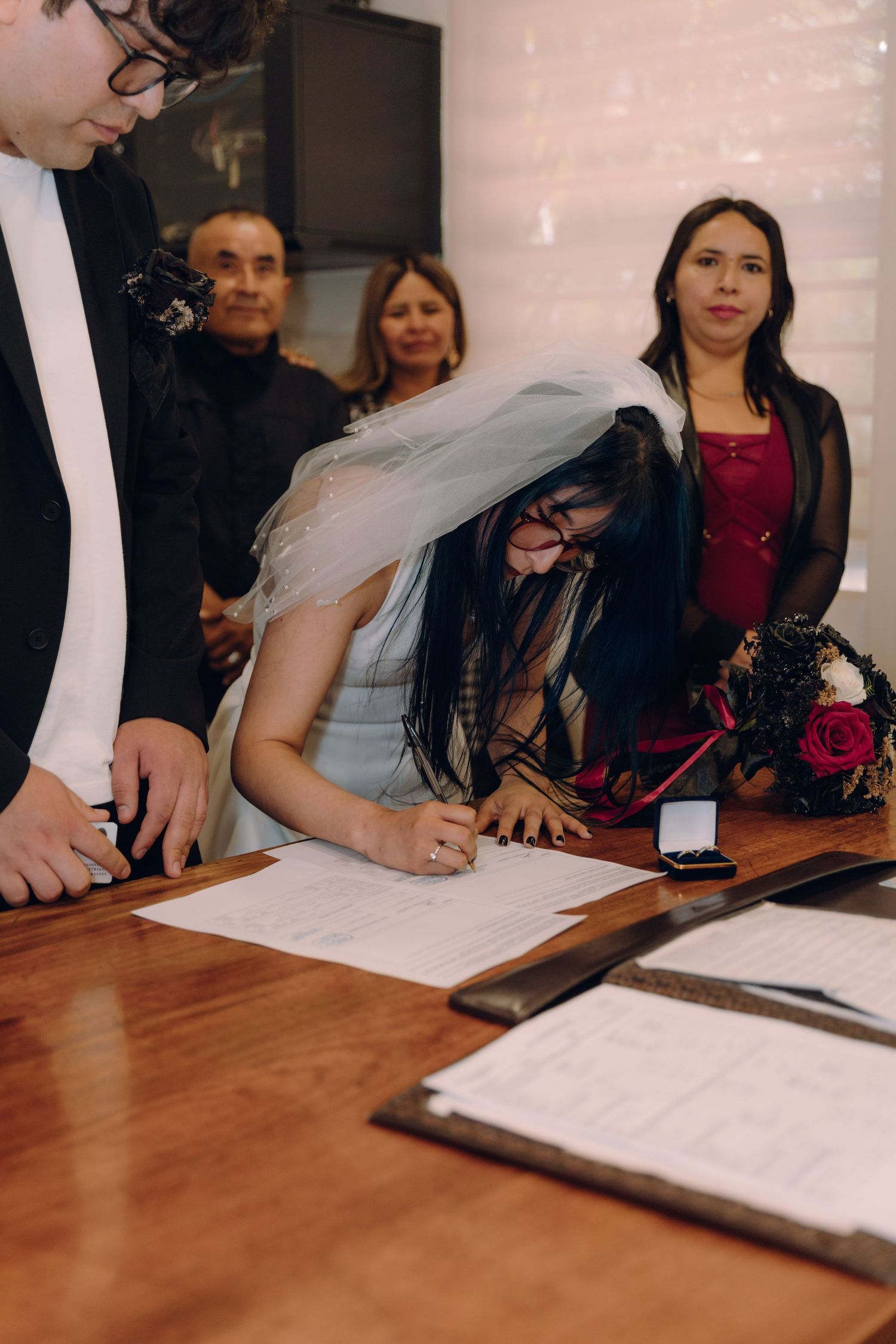 Bride in white dress and veil signing marriage certificate at wooden table with guests watching.