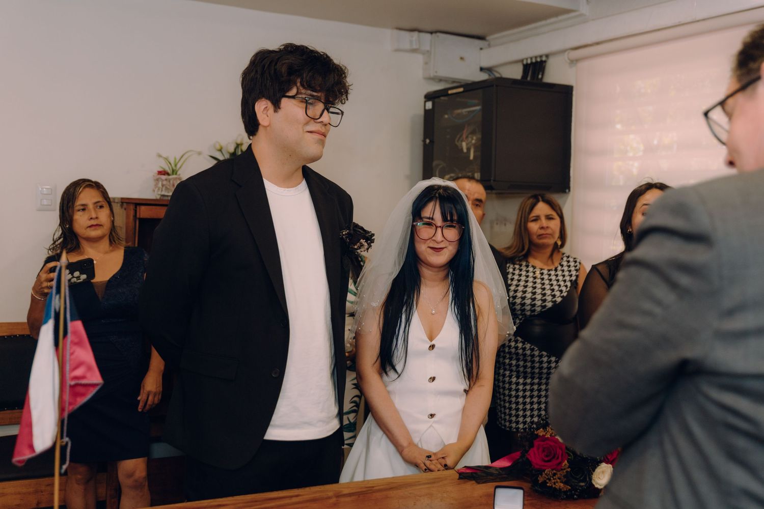 Couple standing before officiant during civil wedding ceremony with smiling guests in background.