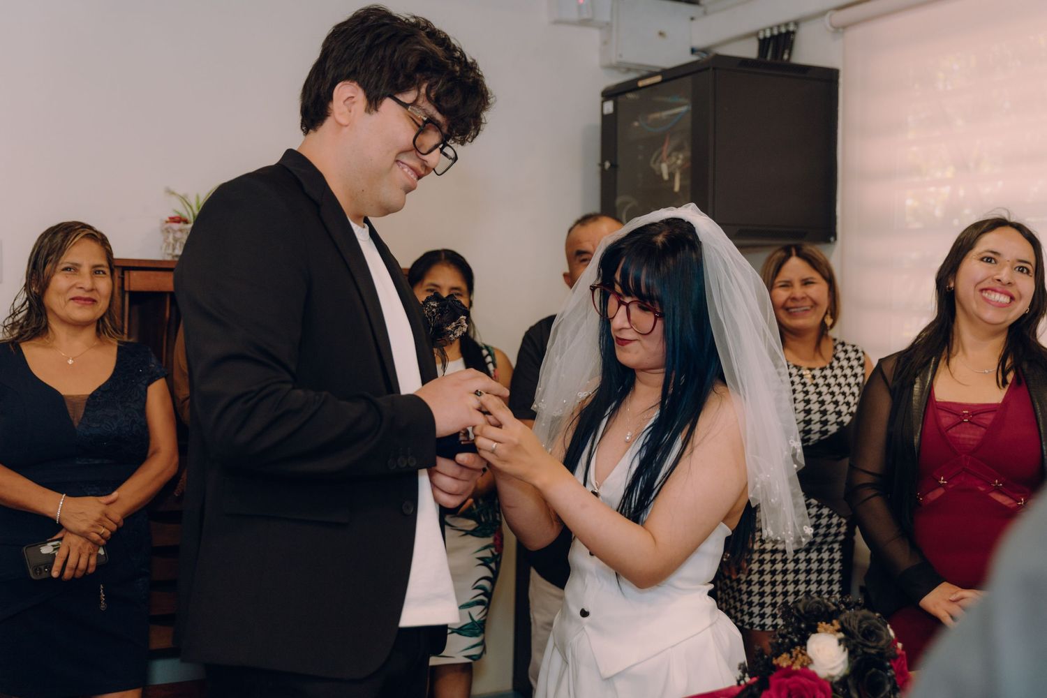 Groom sliding wedding ring onto brides finger as guests smile during civil ceremony.