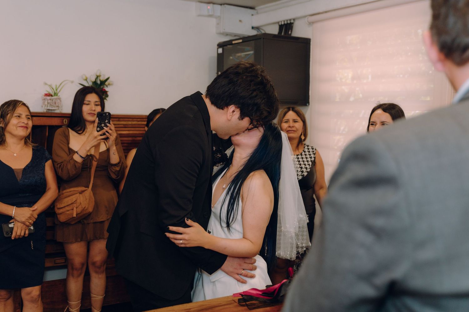 Newlyweds sharing first kiss after civil wedding ceremony with guests watching and photographing.
