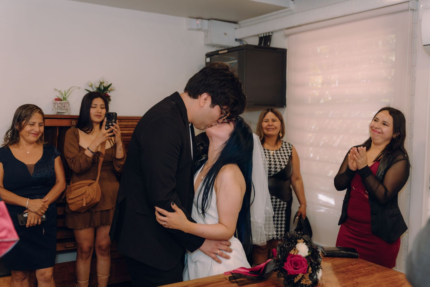 Bride and groom share first kiss at intimate civil wedding ceremony while guests celebrate and take photos.