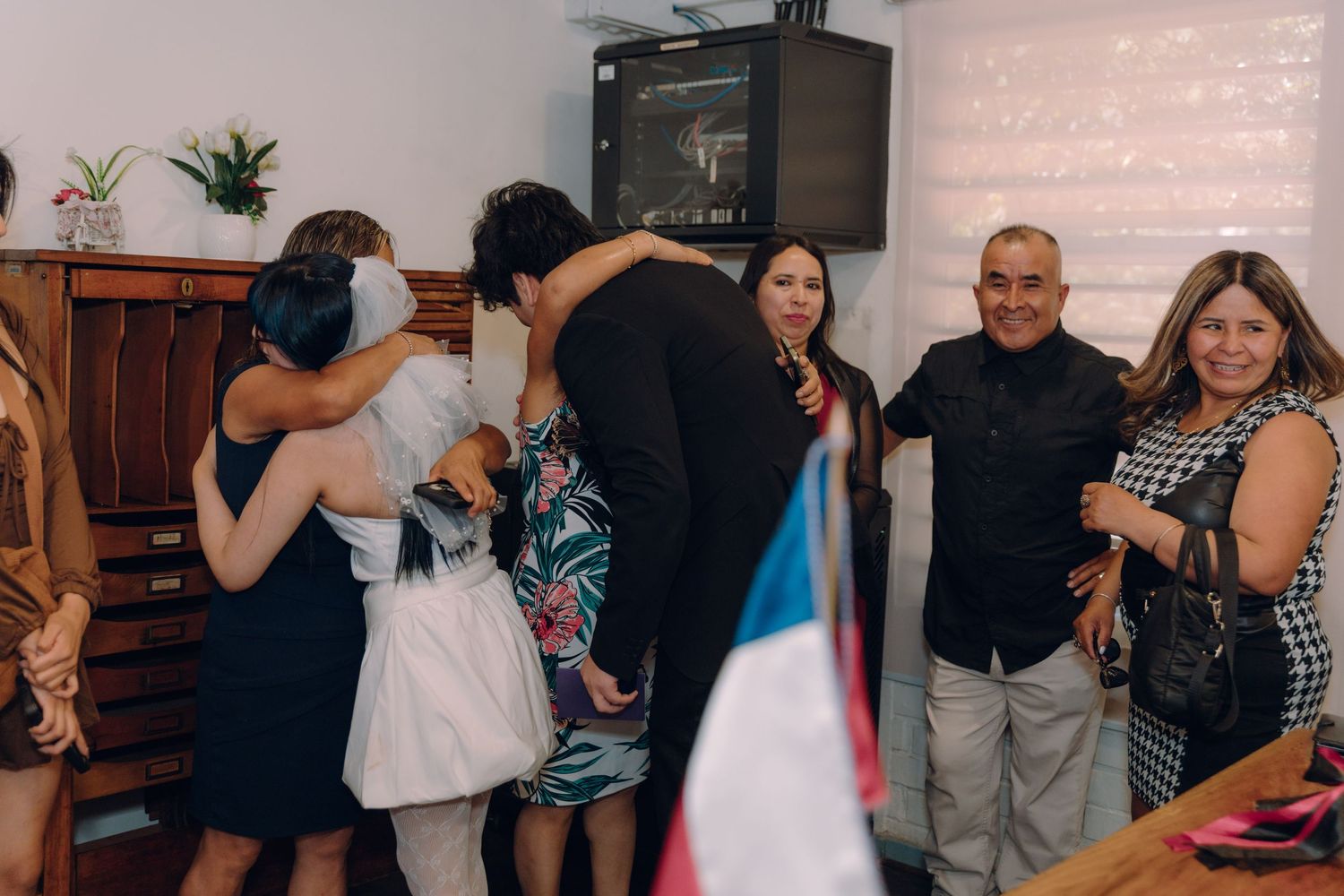 Wedding guests embrace and celebrate with newlyweds at intimate civil ceremony with Chilean flag displayed.